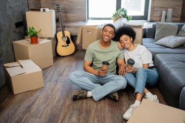 Mutlu Afro-Amerikan çifti yeni evlerinde kutuların yanında otururken ellerinde kahve tutuyorlar.
