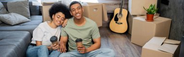 pleased african american couple with takeaway coffee resting near carton boxes in new house, banner clipart