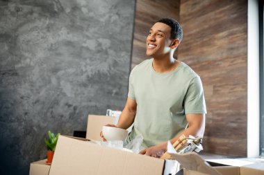 Neşeli Afrikalı Amerikalı adam yeni evdeki paketleri açarken gözlerini kaçırıyor.
