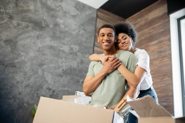 Gülümseyen Afrikalı Amerikalı kadın yeni evdeki paketlerin yanında dururken erkek arkadaşına sarılıyor.