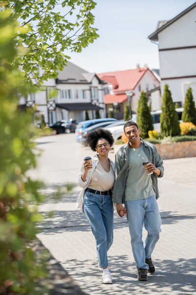 smiling african american couple with coffee to go looking at camera near houses on urban street