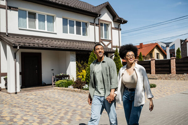 smiling african american couple holding hands while walking together near houses on urban street