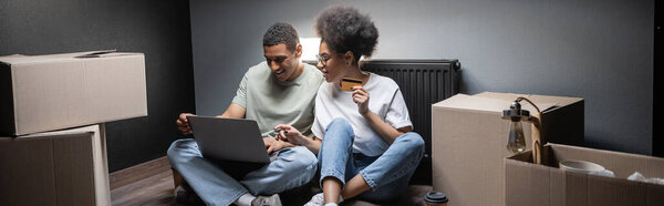 smiling african american couple using laptop and credit card near carton boxes in new house, banner