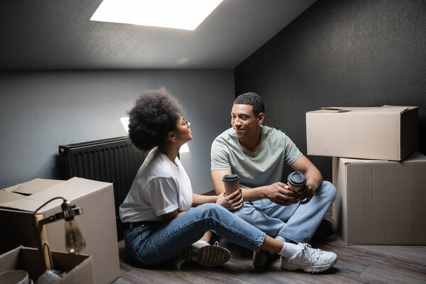 african american woman with coffee to go talking to boyfriend near boxes on attic in new house