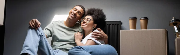 stock image positive african american couple hugging near coffee on carton box on attic in new house, banner