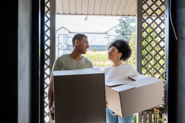 smiling romantic african american couple holding carton boxes near door of new house