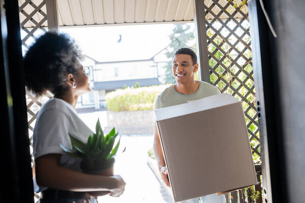 smiling african american man holding carton box near girlfriend with plant in door of new house