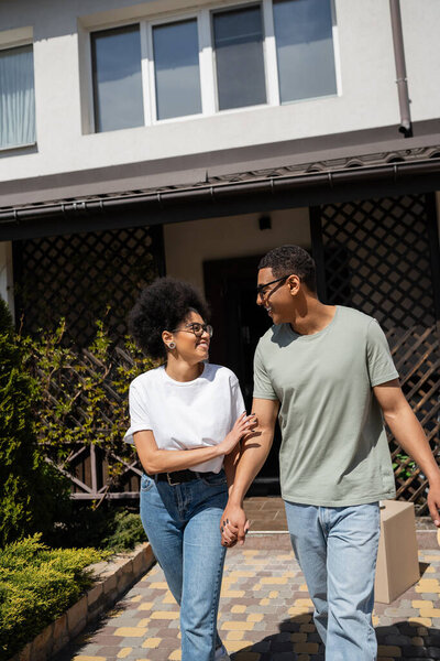 smiling african american woman holding hand of boyfriend in sunglasses near new house