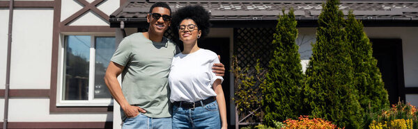 Радостная африканская американская пара обнимается, стоя возле нового дома на открытом воздухе, баннер