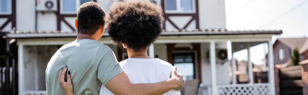 back view of african american couple hugging while standing near new house outdoors, banner