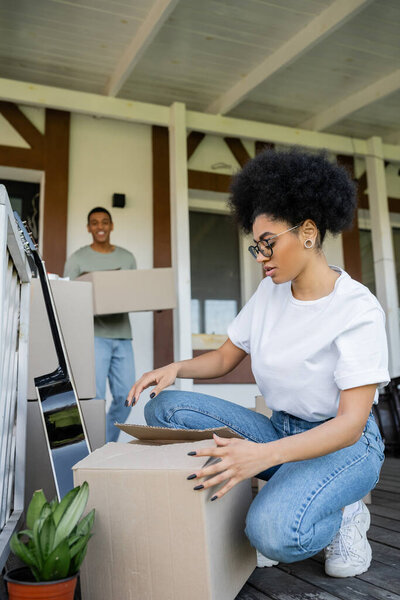african american woman unpacking package near blurred boyfriend, boxes and new house