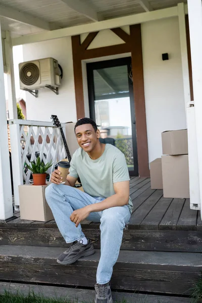 Gülümseyen Afrikalı Amerikalı adam elinde kahve tutarak yeni evin verandasındaki paketlerin yanına gidiyor.