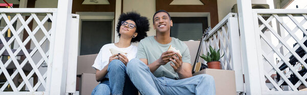 pleased african american couple holding tasty sandwiches on porch of new house, real estate, banner