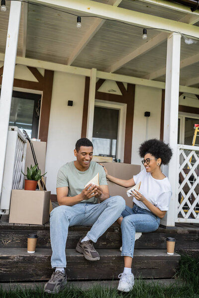 positive african american couple with sandwiches and coffee talking on porch of new house