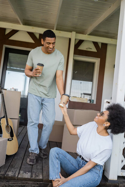 smiling african american man giving coffee to go to girlfriend on porch of new house
