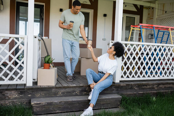 positive african american man giving takeaway coffee to girlfriend near boxes and new house