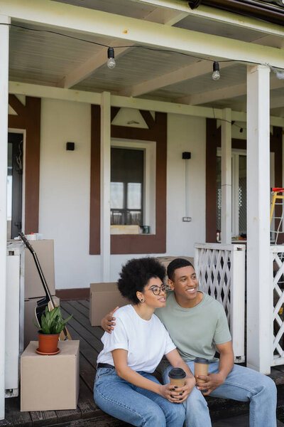 smiling african american man hugging girlfriend with coffee near packages on porch of new house