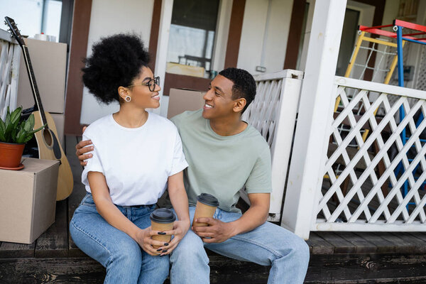 positive african american couple hugging and holding coffee to go near boxes on porch of new house