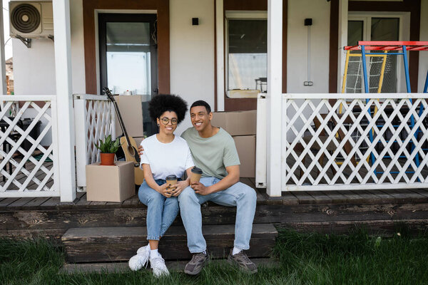 smiling african american couple holding coffee to go near packages on porch of new house