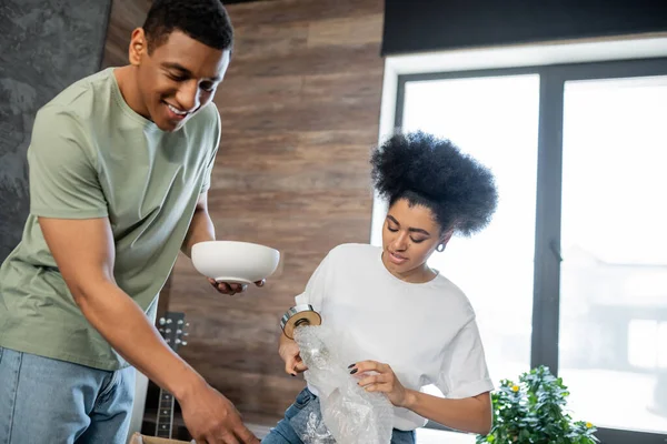 stock image african american woman unpacking floor lamp near boyfriend with tableware in new house