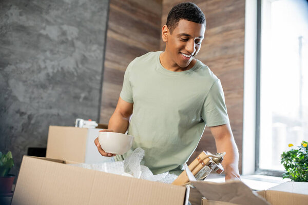 carefree african american man holding belongings near cardboard boxes in new house