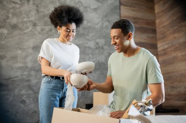 Gülümseyen Afrikalı Amerikalı adam yeni evdeki kutuları açarken kız arkadaşına sofra takımı veriyor.