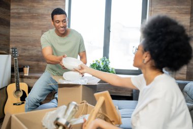 blurred african american woman giving tableware to boyfriend near boxes in new house clipart