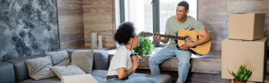 Kahveli mutlu Afro-Amerikan çifti yeni evdeki paketlerin yanında akustik gitar çalıyor.