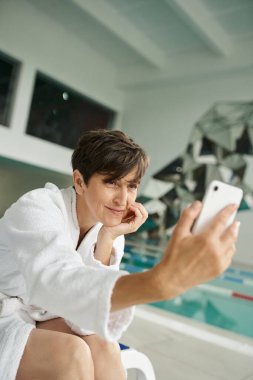 Pozitif orta yaşlı beyaz cüppeli kadın selfie çekiyor, akıllı telefon, spa merkezi, yüzme havuzu.