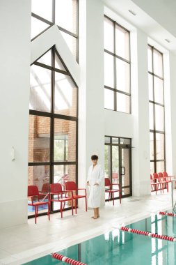 Yüzme havuzlu modern spa merkezi, beyaz cüppeli olgun bir kadın, elleri ceplerinde, rahat bir poz.
