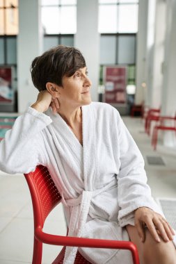 relaxed pose, dreamy mature woman in white robe sitting on red chair, indoor, spa center, look away clipart