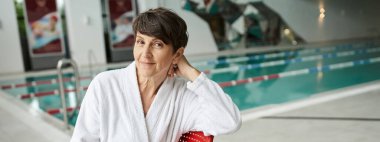 Rahat poz, beyaz bornozlu mutlu olgun bir kadın kapalı havuz kenarında oturuyor, spa günü, pankart