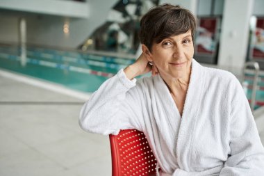 relaxed pose, joyful mature woman in white robe sitting on red chair, indoor swimming pool, spa day clipart