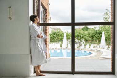 Beyaz cüppeli orta yaşlı bir kadın panoramik pencerenin yanında, havuz merkezinde duruyor.