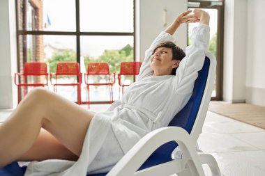middle aged woman with short hair stretching on lounger, white robe, spa center, closed eyes clipart
