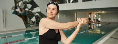 Yüzme şapkalı ve gözlüklü olgun bir kadın yüzme havuzunun, spa merkezinin, mayo ve pankartın yanında ısınıyor.