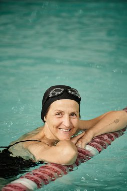 pozitif orta yaşlı kadın havuzda yüzüyor, yüzme şapkası ve gözlükleri takıyor, kameraya, spora, dövmeye bakıyor.