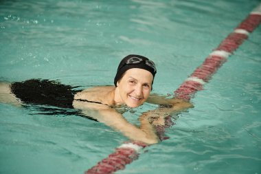 pozitif orta yaşlı bir kadın havuzda yüzüyor, yüzme şapkası ve gözlükleri takıyor, kameraya bakıyor, spor yapıyor.