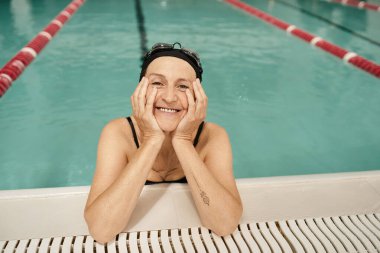 Yüzme şapkası ve gözlüklü neşeli orta yaşlı kadın kameraya bakıyor, havuzdaki su, dinlenme merkezi