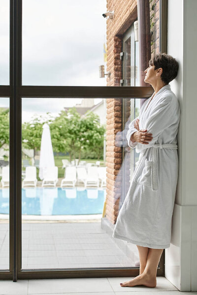mature woman with short hair standing in white robe near panoramic window in spa center, pool view