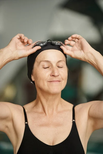 Stock image portrait, active lifestyle, mature woman in swimwear, adjusting goggles, swim cap, spa center