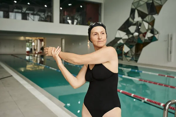 middle aged woman in swim cap and goggles warming up near swimming pool, spa center, swimwear