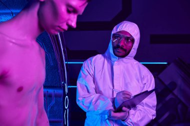 Koruyucu giysi giymiş Hintli bilim adamı panoya yazı yazıyor. Keşif merkezindeki insanımsı uzaylıyı inceliyor.