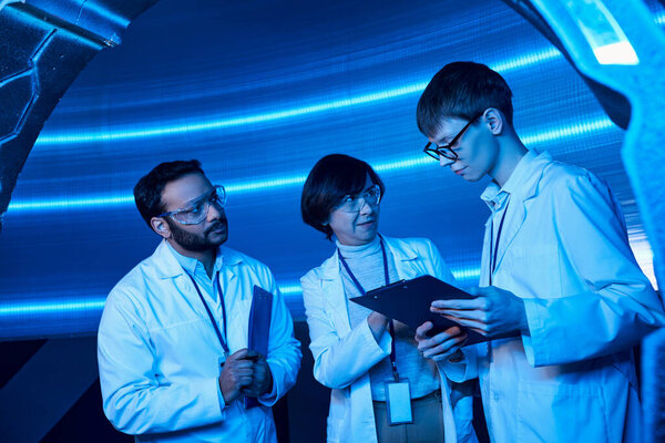 multiethnic scientists looking at young intern with clipboard in neon-lit futuristic science center