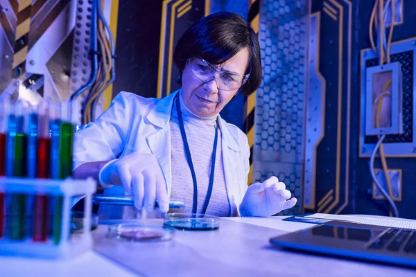 middle aged woman scientist in goggles working with petri dishes and test tubes in futuristic lab
