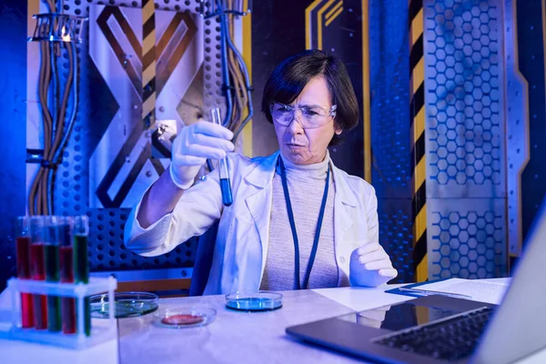 woman scientist in goggles looking at test tube with liquid near laptop in futuristic laboratory