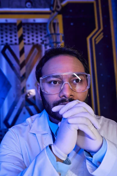 thoughtful indian scientist in goggles and latex gloves in innovation hub, creative thinking
