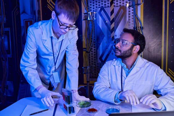 young intern near indian scientist, test tubes with petri dishes in neon-lit futuristic laboratory