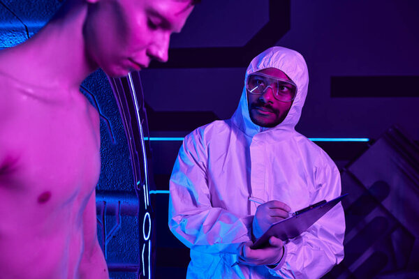 indian scientist in hazmat suit writing on clipboard, examining humanoid alien in discovery center