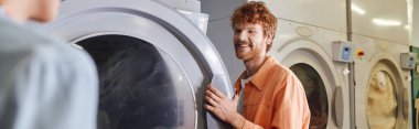 smiling young man standing near washing machines and girlfriend in self service laundry, banner clipart
